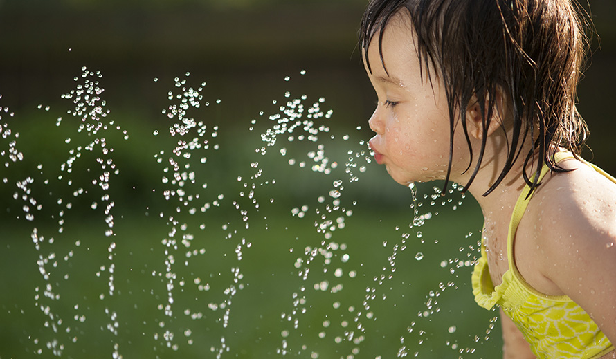 Una niña pequeña se refresca bebiendo de un rociador. 