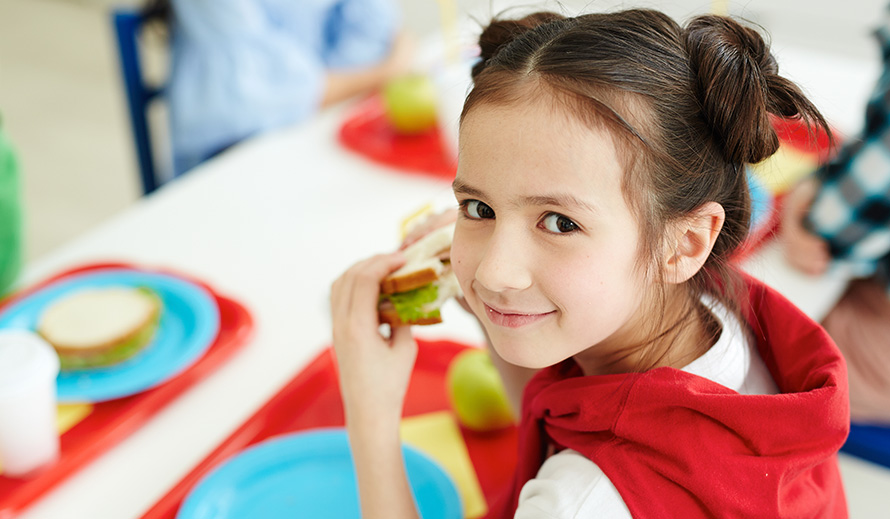 A little girl smiles while eating school lunch. 