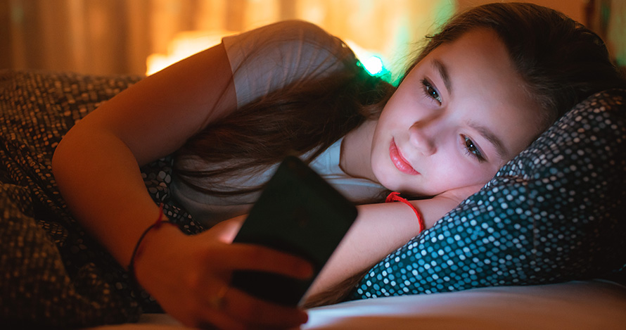 A young Hispanic girl stares at her phone while in bed. 