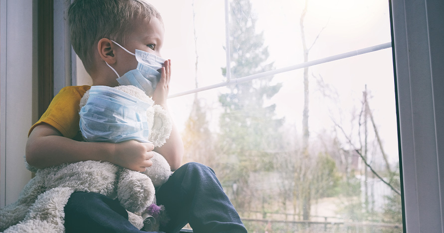 Young boy with a mask sits looking out his window during quarantine.