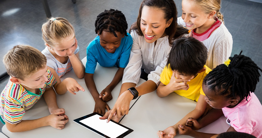 An early learning teacher talks to a group of children surrounding her at a table. 