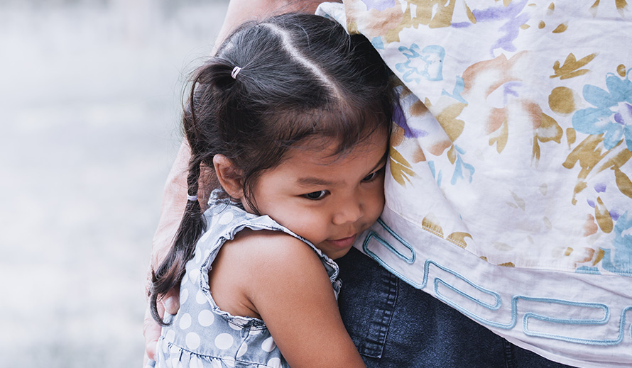 A little Hispanic girl, crying and standing next to her mother with her arms wrapped around her waist.