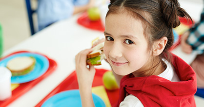 A little girl smiles while eating school lunch. 