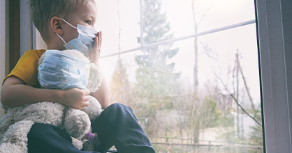 Young boy with a mask sits looking out his window during quarantine.