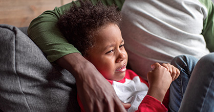 A distressed young African-American boy curled up on the couch being comforted by his father.