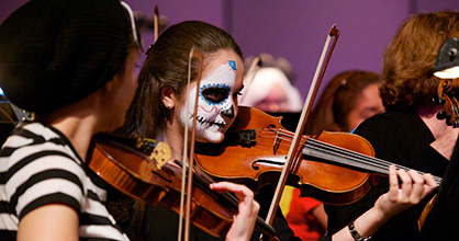 A costumed symphony orchestra performing a concert while cartoon ghosts, bats and the words “Spooky Symphony” are projected onto a big screen above them.